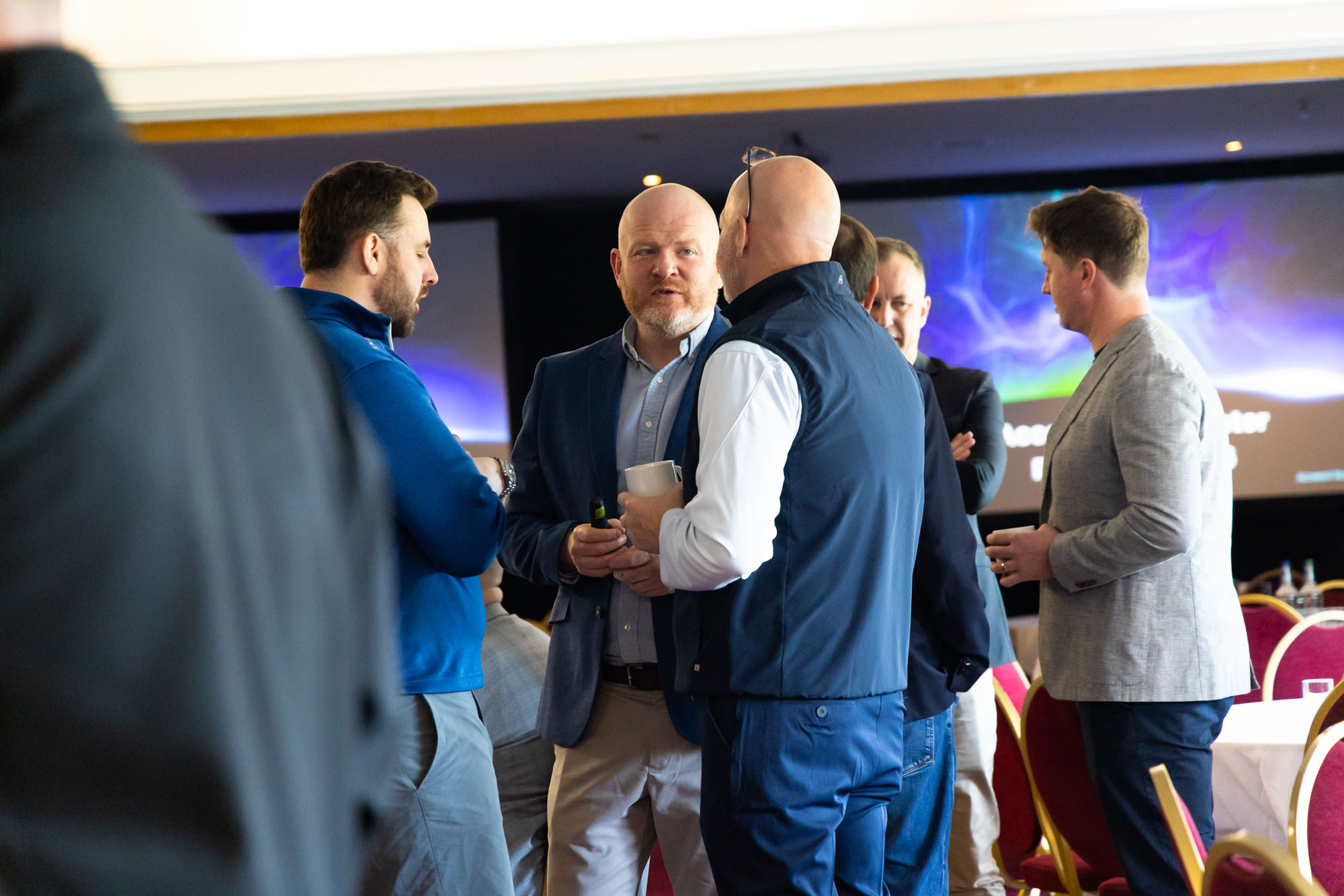 Group of men in smart-casual attire chat and hold drinks at an indoor networking event or reception.
