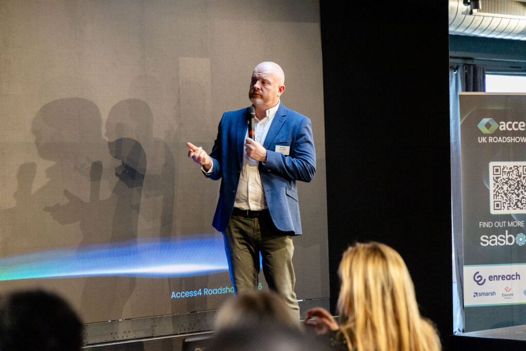 Speaker on stage delivering a talk, holding a microphone, wearing a blue blazer and white shirt, with an audience in front and banners to the right.