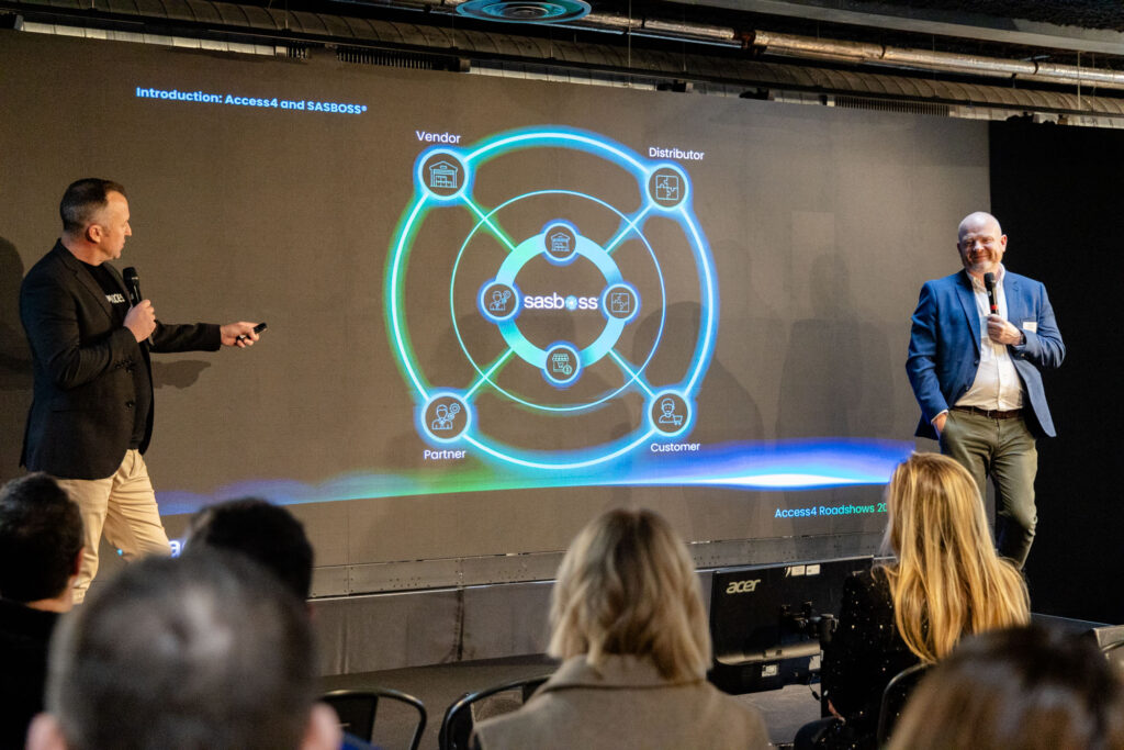 Two men on a stage presenting a large circular diagram projected on a screen, with the sasboss logo at center and labels like Vendor, Distributor, Partner, and Customer around the rings.