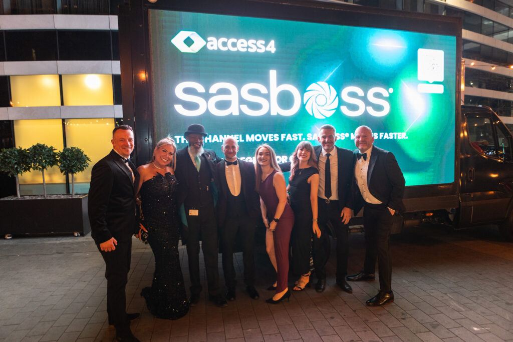 Nine adults in formal evening wear pose for a group photo in front of a large, bright digital billboard at night (access4 logo visible).