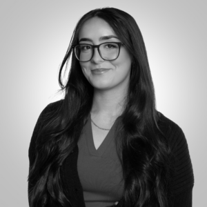 Smiling woman with long dark hair and glasses, wearing a V-neck top and cardigan; head-and-shoulders portrait against a light gradient background.