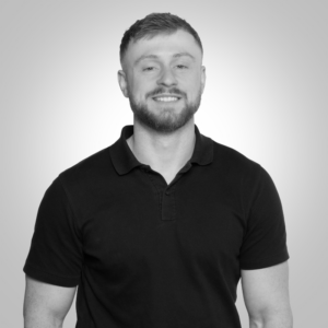 Smiling man with short hair and a beard, wearing a black polo shirt, posed for a head-and-shoulders portrait against a gray gradient background.