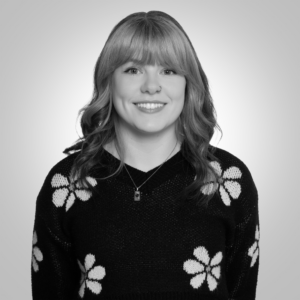 Portrait of a smiling woman with shoulder-length hair wearing a black sweater with white flower patterns, against a gray gradient background.
