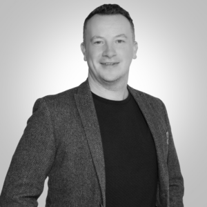 Professional headshot of a smiling man in a gray herringbone blazer over a black shirt, facing the camera.