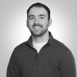 Portrait of a smiling man with short dark hair and a light beard wearing a dark quarter-zip sweater against a neutral gray background.