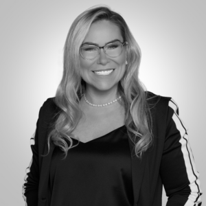 Portrait of a smiling woman wearing glasses, a black blazer with white striped sleeves, and a pearl necklace.
