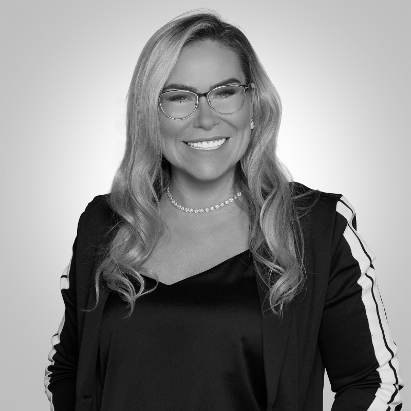 Portrait of a smiling woman wearing glasses, a black blazer with white striped sleeves, and a pearl necklace.