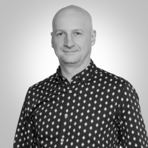Head-and-shoulders portrait of a bald man wearing a dark shirt with a white geometric pattern, neutral studio background, slight smile.