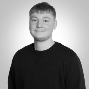 Portrait of a young man with short hair wearing a black crewneck sweater, facing the camera with a subtle smile, against a light gray background.